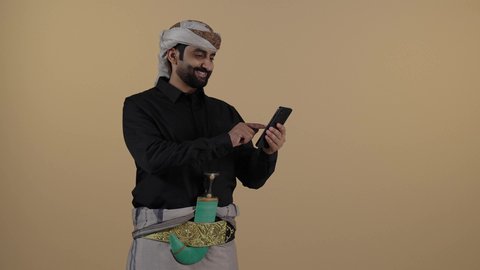 بورتريه رجل عربي يمني يرتدي القميص والازار مع العمامة والجنبية يقف ويحمل هاتف، مواقع التواصل الاجتماعي والتصفح، مفهوم لغة الجسد وتعابير الوجه، تمثيل الرجولة العربية الأصيلة، الاجهزة التقنية الحديثة في الحياة اليومية، مفهوم الهيبة والاناقة، خلفية بيج