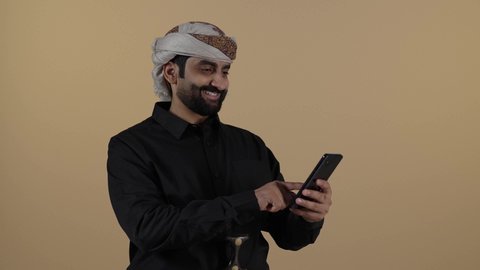 بورتريه رجل عربي يمني يرتدي القميص والازار مع العمامة  والجنبية يقف ويحمل هاتف، استخدام الاجهزة الحديثة في التواصل، مفهوم لغة الجسد وتعابيرالوجه، تمثيل الرجولة العربية الأصيلة، الاجهزة التقنية الحديثة في الحياة اليومية، مفهوم الهيبة والاناقة، خلفية بيج