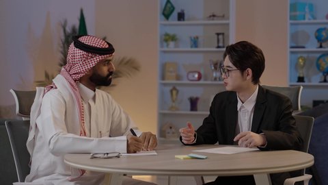 اعداد تقارير العمل، تبادل الخبرات والمعرفة بين البلاد، استضافة رجال الاعمال الاجانب في المملكة العربية السعودية، رجل عربي خليجي سعودي يرتدي الزي التقليدي مع شاب صيني يرتدي البدلة الرسمية يقومان بمناقشة المشاريع، المناقشة في اهداف الشركة