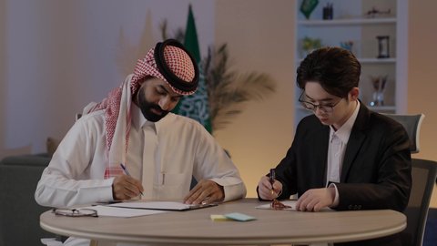 Preparing work reports, exchanging experiences and knowledge between countries, hosting foreign businessmen in the Kingdom of Saudi Arabia, an Arab Gulf Saudi man wearing traditional attire with a Chinese young man in a formal suit discussing projects, discussing the company's goals.