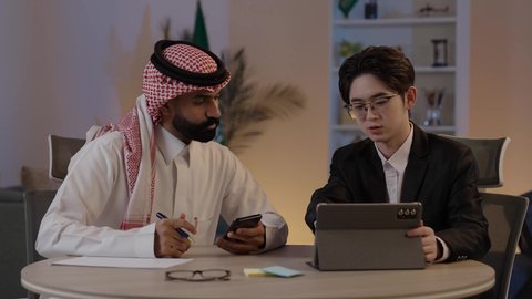 Exchange of experiences and knowledge between countries, discussion of business and projects, hosting foreign businessmen in the Kingdom, using modern technology to accomplish work, an Arab Gulf Saudi man wearing traditional attire with a Chinese young man in a formal suit viewing a project on a mobile tablet.