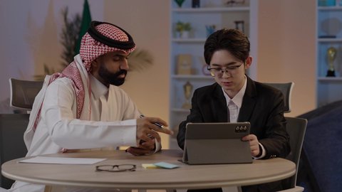 Discussion on business and projects, hosting foreign businessmen in the Kingdom, using modern technology to accomplish work, exchanging experiences and knowledge between countries, an Arab Gulf Saudi man wearing traditional attire with a Chinese young man in a formal suit viewing a project on a mobile tablet.