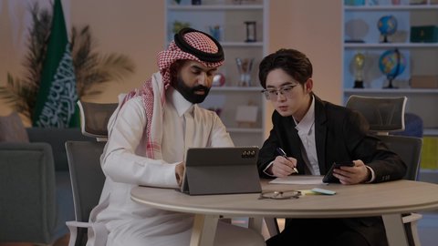 Exchange of experiences and knowledge between countries, discussion of business and projects, hosting foreign businessmen in the Kingdom, using modern technology to accomplish work, an Arab Gulf Saudi man wearing traditional attire with a Chinese young man in a formal suit viewing a project on a mobile tablet.
