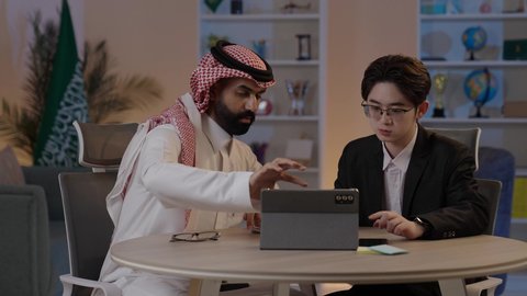 المناقشة في الاعمال والمشاريع، استضافة رجال الاعمال الاجانب في المملكة, استخدام التقنية الحديثة في انجاز العمل، تبادل الخبرات والمعرفة بين البلاد, رجل عربي خليجي سعودي يرتدي الزي التقليدي مع شاب صيني يرتدي البدلة الرسمية يقومان برؤية مشروع ما على التابليت المحمول