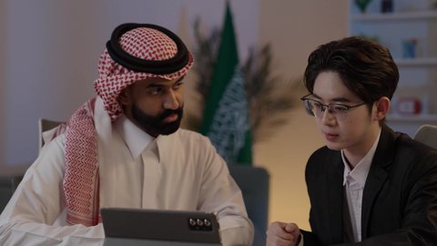 Exchange of experiences and knowledge between countries, discussion of business and projects, hosting foreign businessmen in the Kingdom, using modern technology to accomplish work, an Arab Gulf Saudi man wearing traditional attire with a Chinese young man in a formal suit viewing a project on a mobile tablet.