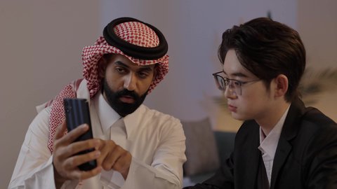 تبادل الخبرات والمعرفة بين البلاد, المناقشة في الاعمال والمشاريع، استضافة رجال الاعمال الاجانب في المملكة, استخدام التقنية الحديثة في انجاز العمل، رجل عربي خليجي سعودي يرتدي الزي التقليدي مع شاب صيني يرتدي البدلة الرسمية يقومان برؤية مشروع ما على الهاتف المحمول