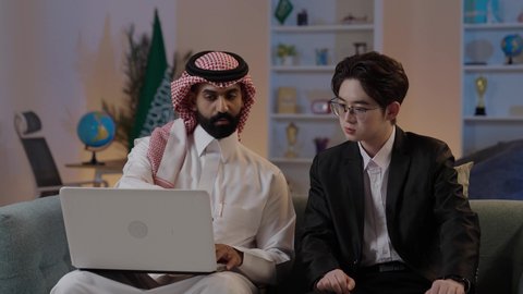 استضافة رجال الاعمال الاجانب في المملكة, استخدام التقنية الحديثة في انجاز العمل، تبادل الخبرات والمعرفة بين البلاد, عقد اتفاقيات عمل جديدة، رجل عربي خليجي سعودي يرتدي الزي التقليدي مع شاب صيني يرتدي البدلة الرسمية يقومان برؤية مشروع ما على اللابتوب المحمول