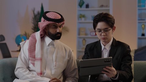 استخدام التقنية الحديثة في انجاز العمل، تبادل الخبرات والمعرفة بين البلاد, عقد اتفاقيات عمل جديدة، استضافة رجال الاعمال الاجانب في المملكة, رجل عربي خليجي سعودي يرتدي الزي التقليدي مع شاب صيني يرتدي البدلة الرسمية يقومان برؤية مشروع ما على التابليت المحمول