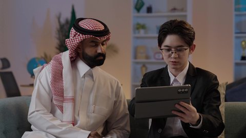 استضافة رجال الاعمال الاجانب في المملكة, استخدام التقنية الحديثة في انجاز العمل، تبادل الخبرات والمعرفة بين البلاد, عقد اتفاقيات عمل جديدة، رجل عربي خليجي سعودي يرتدي الزي التقليدي مع شاب صيني يرتدي البدلة الرسمية يقومان برؤية مشروع ما على التابليت المحمول