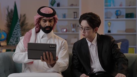 استخدام التقنية الحديثة في انجاز العمل، تبادل الخبرات والمعرفة بين البلاد, عقد اتفاقيات عمل جديدة، استضافة رجال الاعمال الاجانب في المملكة, رجل عربي خليجي سعودي يرتدي الزي التقليدي مع شاب صيني يرتدي البدلة الرسمية يقومان برؤية مشروع ما على التابليت المحمول