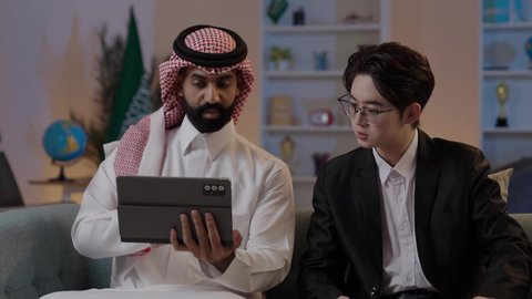 استضافة رجال الاعمال الاجانب في المملكة, استخدام التقنية الحديثة في انجاز العمل، تبادل الخبرات والمعرفة بين البلاد, عقد اتفاقيات عمل جديدة، رجل عربي خليجي سعودي يرتدي الزي التقليدي مع شاب صيني يرتدي البدلة الرسمية يقومان برؤية مشروع ما على التابليت المحمول
