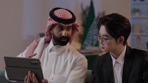 استضافة رجال الاعمال الاجانب في المملكة, استخدام التقنية الحديثة في انجاز العمل، تبادل الخبرات والمعرفة بين البلاد, عقد اتفاقيات عمل جديدة، رجل عربي خليجي سعودي يرتدي الزي التقليدي مع شاب صيني يرتدي البدلة الرسمية يقومان برؤية مشروع ما على التابليت المحمول