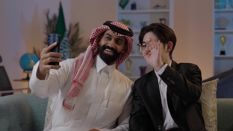The meeting of greetings and peace with gestures of joy, the exchange of experiences and knowledge between countries, hosting foreign businessmen in the Kingdom, an Arab Gulf Saudi man wearing traditional attire with a Chinese young man in a formal suit conducting a video call via mobile phone.