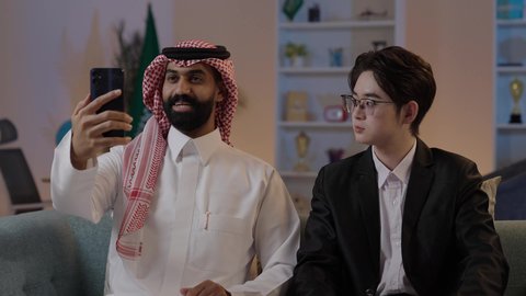 التقاء التحية والسلام بإيماءات السرور، تبادل الخبرات والمعرفة بين البلاد، استضافة رجال الاعمال الاجانب في المملكة، رجل عربي خليجي سعودي يرتدي الزي التقليدي مع شاب صيني يرتدي البدلة الرسمية يقومان بإجراء مكالمة فيديو عبر الهاتف المحمول