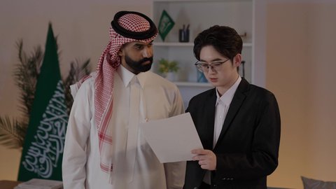 Preparing work reports, exchanging experiences and knowledge between countries, hosting foreign businessmen in the Kingdom of Saudi Arabia, an Arab Gulf Saudi man wearing traditional attire with a Chinese young man in formal suit discussing projects, discussing the company's goals.