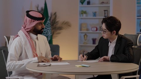 اعداد تقارير العمل، تبادل الخبرات والمعرفة بين البلاد، استضافة رجال الاعمال الاجانب في المملكة العربية السعودية، رجل عربي خليجي سعودي يرتدي الزي التقليدي مع شاب صيني يرتدي البدلة الرسمية يقومان بمناقشة المشاريع، المناقشة في اهداف الشركة
