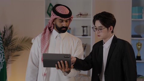 Signing new work agreements, hosting foreign businessmen in the Kingdom of Saudi Arabia, exchanging experiences and knowledge between countries, an Arab Gulf Saudi man wearing traditional attire with a Chinese young man in a formal suit working on a tablet.