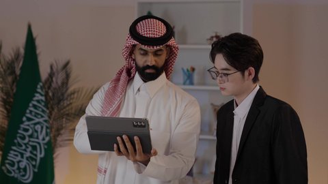 Exchange of experiences and knowledge between countries, signing new business agreements, hosting foreign businessmen in the Kingdom of Saudi Arabia, an Arab Gulf Saudi man wearing traditional attire with a Chinese young man in a formal suit working on a mobile tablet.