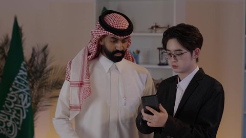 Exchange of experiences and knowledge between countries, signing new business agreements, hosting foreign businessmen in the Kingdom of Saudi Arabia, an Arab Gulf Saudi man wearing traditional attire with a Chinese young man in a formal suit looking at a project on a mobile phone.