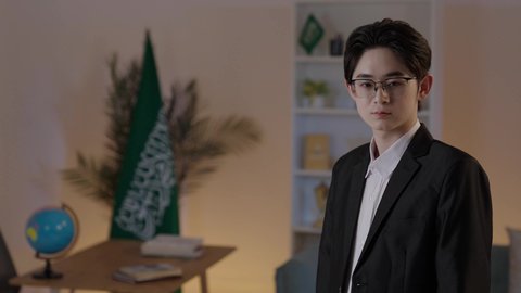 Standing with crossed arms and looking at the camera with gestures of happiness and joy, hosting foreign businessmen in the Kingdom of Saudi Arabia, a young Chinese man wearing a formal suit and glasses, paying attention to external appearance.