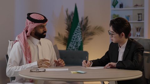 Hosting foreign businessmen in the Kingdom of Saudi Arabia, exchanging experiences and knowledge between countries, a portrait of an Arab Gulf Saudi man wearing traditional attire with a Chinese young man in a formal suit working on a laptop.