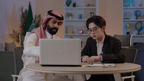 عقد اتفاقيات عمل جديدة، استضافة رجال الاعمال الاجانب في المملكة العربية السعودية, تبادل الخبرات والمعرفة بين البلاد, رجل عربي خليجي سعودي يرتدي الزي التقليدي مع شاب صيني يرتدي البدلة الرسمية يقومان بالعمل على اللابتوب المحمول