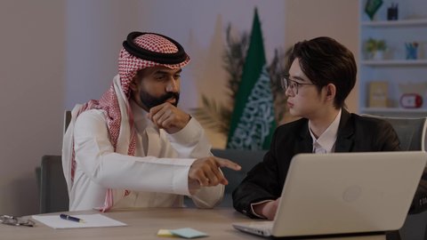 عقد اتفاقيات عمل جديدة، استضافة رجال الاعمال الاجانب في المملكة العربية السعودية, تبادل الخبرات والمعرفة بين البلاد, رجل عربي خليجي سعودي يرتدي الزي التقليدي مع شاب صيني يرتدي البدلة الرسمية يقومان بالعمل على اللابتوب المحمول