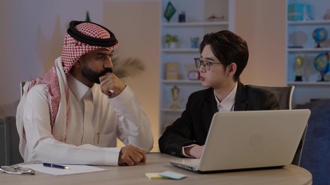 استضافة رجال الاعمال الاجانب في المملكة العربية السعودية, تبادل الخبرات والمعرفة بين البلاد, بورتريه لرجل عربي خليجي سعودي يرتدي الزي التقليدي مع شاب صيني يرتدي البدلة الرسمية يقومان بالعمل على اللابتوب المحمول