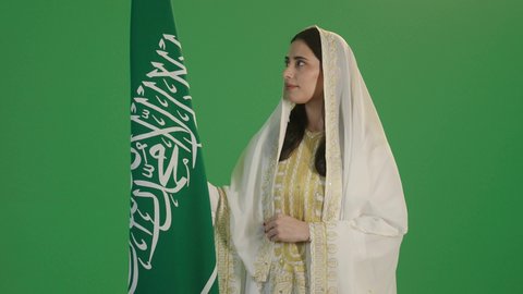 الأزياء الشعبية التقليدية، اليوم الوطني السعودي 23 سبتمبر، يوم العلم 11 مارس، بورتريه مقرب لامرأة عربية خليجية سعودية مبتسمة ترتدي الشيلة والنشل تمسك علم المملكة بإيماءات الفخر، ارتداء زي المنطقة الشرقية، خلفية خضراء, كروما