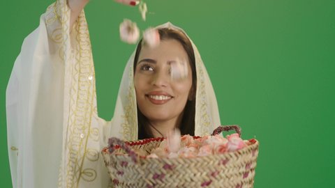 A portrait of a young Arab Gulf Saudi woman wearing a white dress embroidered with gold and wearing a shawl on her head, holding a basket made of straw filled with natural damask roses in her hand. This is the traditional Saudi folk attire, suitable for national occasions and holidays, commemorating the founding of the first Saudi state on February 22, with a green background, chroma.