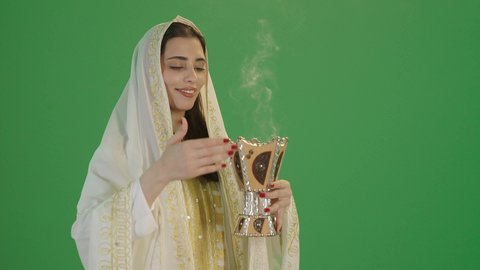 استخدام خشب العود بالأعياد والمناسبات,ارتداء الزي السعودي التقليدي الشعبي, بورتريه لإمرأة عربية خليجية سعودية ترتدي الثوب الابيض المطرز بالذهبي وتضع الشيله على رأسها تمسك المبخرة التراثية المزخرفة بيدها و تنظر إلى الكاميرا بإيماءات السعادة و السرور, مفهوم التعطر بالبخور, خلفية خضراء, كروما
