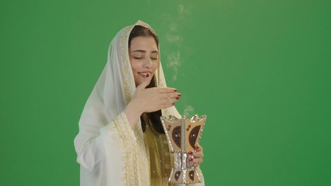مفهوم التعطر بالبخور, استخدام خشب العود بالأعياد والمناسبات, ارتداء الزي السعودي التقليدي الشعبي, بورتريه لإمرأة عربية خليجية سعودية ترتدي الثوب الابيض المطرز بالذهبي وتضع الشيله على رأسها تمسك المبخرة التراثية المزخرفة بيدها و تنظر إلى الكاميرا بإيماءات السعادة و السرور, خلفية خضراء, كروما