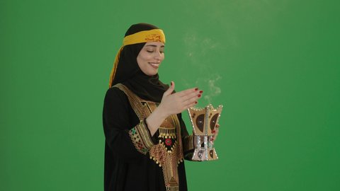 عادات وتقاليد المجتمع السعودي في الأعياد والمناسبات الاسلامية, بورتريه من الجانب  لإمرأة عربية خليجية سعودية مبتسمة ترتدي الكرتة والهامة تمسك المبخرة التراثية المزخرفة بيدها و تنظر إلى الكاميرا بإيماءات السعادة و السرور, خلفية خضراء, كروما
