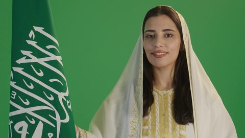 السعادة بالانتماء للهوية السعودية، الأزياء الشعبية التقليدية، اليوم الوطني السعودي 23 سبتمبر، يوم العلم 11 مارس، بورتريه مقرب لامرأة عربية خليجية سعودية مبتسمة ترتدي الشيلة والنشل تمسك علم المملكة بإيماءات الفخر، ارتداء زي المنطقة الشرقية، خلفية خضراء, كروما