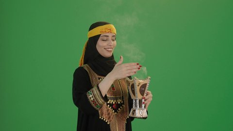 مفهوم التعطر بالبخور, بورتريه من الجانب لإمرأة عربية خليجية سعودية مبتسمة ترتدي الكرتة والهامة, تمسك المبخرة التراثية المزخرفة بيدها و تنظر إلى الكاميرا بإيماءات السعادة و السرور, خلفية خضراء, كروما