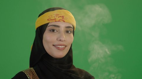 استخدام خشب العود بالأعياد والمناسبات, بورتريه مقرب لإمرأة عربية خليجية سعودية مبتسمة ترتدي الكرتة والهامة, تمسك المبخرة التراثية المزخرفة بيدها و تنظر إلى الكاميرا بإيماءات السعادة و السرور, خلفية خضراء, كروما