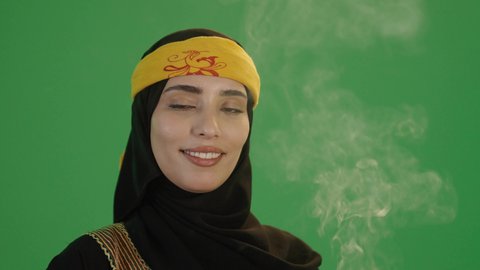 التعطر و التطيب باطيب الروائح والعطور, استخدام خشب العود بالأعياد والمناسبات, بورتريه مقرب لإمرأة عربية خليجية سعودية مبتسمة ترتدي الكرتة والهامة, تمسك المبخرة التراثية المزخرفة بيدها و تنظر إلى الكاميرا بإيماءات السعادة و السرور, خلفية خضراء, كروما