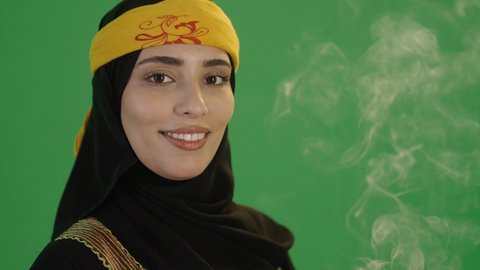 إيماءات السعادة و السرور, عادات وتقاليد المجتمع السعودي في الأعياد والمناسبات الاسلامية, بورتريه مقرب لإمرأة عربية خليجية سعودية مبتسمة ترتدي الكرتة والهامة, تمسك المبخرة التراثية المزخرفة بيدها وتنظر إلى الكاميرا , خلفية خضراء، كروما