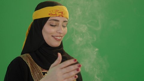 العراقة و الاصالة  والتراث السعودي, استخدام خشب العود بالأعياد والمناسبات, بورتريه لإمرأة عربية خليجية سعودية مبتسمة ترتدي الكرتة والهامة, تمسك المبخرة التراثية المزخرفة بيدها و تنظر إلى الكاميرا بإيماءات السعادة و السرور, مفهوم التعطر بالبخور, خلفية خضراء، كروما