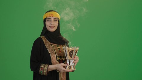 أصالة العادات والتقاليد الخليجية, استخدام خشب العود بالأعياد والمناسبات, بورتريه لإمرأة عربية خليجية سعودية مبتسمة ترتدي الكرتة والهامة, تمسك المبخرة التراثية المزخرفة بيدها و تنظر إلى الكاميرا بإيماءات السعادة و السرور, مفهوم التعطر بالبخور, خلفية خضراء, كروما
