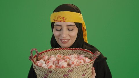 مفهوم حب الوطن والانتماء اليه، المناسبات والأعياد الوطنية، ذكرى تأسيس الدولة السعودية الاولى 22 فبراير، بورتريه مقرب لإمرأة عربية خليجية سعودية ترتدي الكرتة والهامة تنظرالى الكاميرا تحمل بيدها سلة من القش ممتلئة بالورد الجوري الطبيعي، يوم بدينا 1727 م، خلفية خضراء، كروما