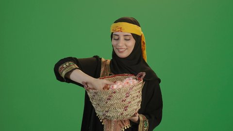 ذكرى تأسيس الدولة السعودية الاولى 22 فبراير، المناسبات والأعياد الوطنية، بورتريه مقرب لإمرأة عربية خليجية سعودية ترتدي الكرتة والهامة تنظرالى الكاميرا تحمل بيدها سلة من القش ممتلئة بالورد الجوري الطبيعي، يوم بدينا 1727 م، خلفية خضراء، كروما