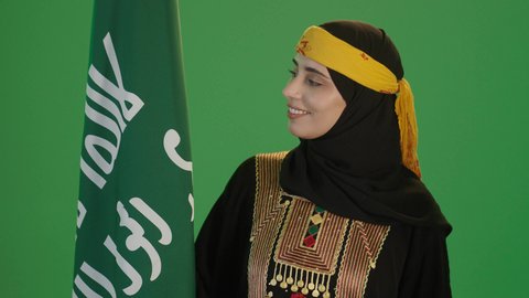 ارتداء زي المنطقة الوسطى، النظر إلى العلم بفخر، الاحتفال بالمناسبات الوطنية، علم يوم تأسيس الدولة السعودية, يوم بدينا 1727م, الفخر بالتراث التاريخي، يوم العلم 11 مارس، بورتريه مقرب لإمرأة عربية خليجية سعودية ترتدي الكرتة والهامة تحمل علم المملكة, خلفية خضراء، كروما
