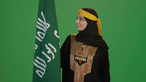 يوم بدينا 1727 م، ذكرى تأسيس الدولة السعودية الأولى 22 فبراير، ارتداء أزياء المنطقة الوسطى، بورتريه لإمرأة عربية خليجية سعودية مبتسمة ترتدي الكرتة والهامة تنظر إلى علم يوم التأسيس بفخر، الاحتفال بالمناسبات والأعياد الوطنية الشعبية، خلفية خضراء، كروما