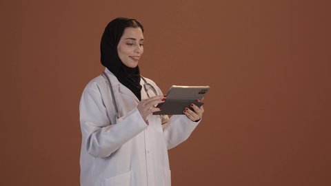 وصف العلاج والتوعيه الطبيه والتثقيفيه, متابعة وتدوين معلومات السجل الطبي الالكتروني الخاص بالمريض, بورتريه من الجانب لطبيبة عربيه خليجيه سعودية محجبة ترتدي اليونيفورم الطبي تحمل بيدها جهاز التابلت، ، خلفيه بنية