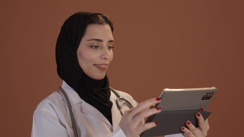 متابعة وتدوين معلومات السجل الطبي الالكتروني الخاص بالمريض, بورتريه من الجانب لطبيبة عربيه خليجيه سعودية محجبة ترتدي اليونيفورم الطبي تحمل بيدها جهاز التابلت، وصف العلاج والتوعيه الطبيه والتثقيفيه، خلفيه بنية