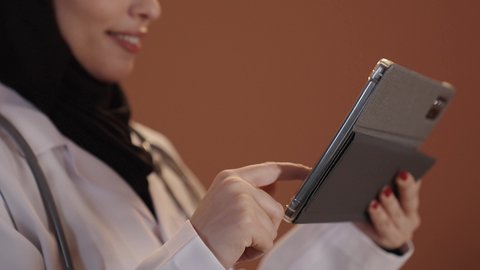 اجراء الاستشارات الطبية عبر الأونلاين, تصويرمقرب لطبيبة عربية خليجية سعودية محجبة ترتدي المعطف الطبي تعمل على جهاز التابليت, تشخيص الحالات المرضية وتوفير الخدمات للمرضى, استخدام الأجهزة والتقنيات الحديثة في البحث الطبي, خلفية بنية