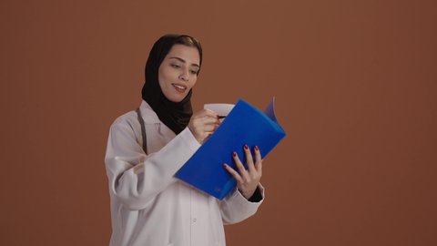 قراءة نتائج الفحوصات بالسجل الطبي, مفهوم الطب و الرعاية الصحية، بورتريه مقرب لطبيبة عربية خليجية سعودية محجبة مبتسمه ترتدي المعطف مع السماعه الطبيه تقوم بتسجيل معلومات المرضى, تقديم الخدمات العلاجية في المراكز الطبية, خلفيه بنية
