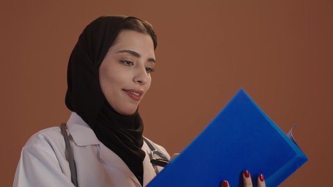 كتابة وتدوين التقارير والحالات المرضية, مفهوم الطب و الرعاية الصحية، بورتريه مقرب لطبيبة عربية خليجية سعودية محجبة مبتسمه ترتدي المعطف مع السماعه الطبيه تقوم بتسجيل معلومات المرضى, تقديم الخدمات العلاجية في المراكز الطبية, خلفيه بنية