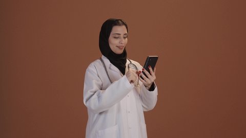 تقديم الخدمات للمرضى بكل كفاءه, إجراء الاستشارت الطبية والخدمات العلاجية للمرضى عبر الأونلاين, استخدام الجوال في البحث والتطوير، بورتريه مقرب لطبيبة عربية خليجية سعودية محجبة ترتدي المعطف الابيض وتضع سماعتها الطبية حول رقبتها، خلفيه بنية